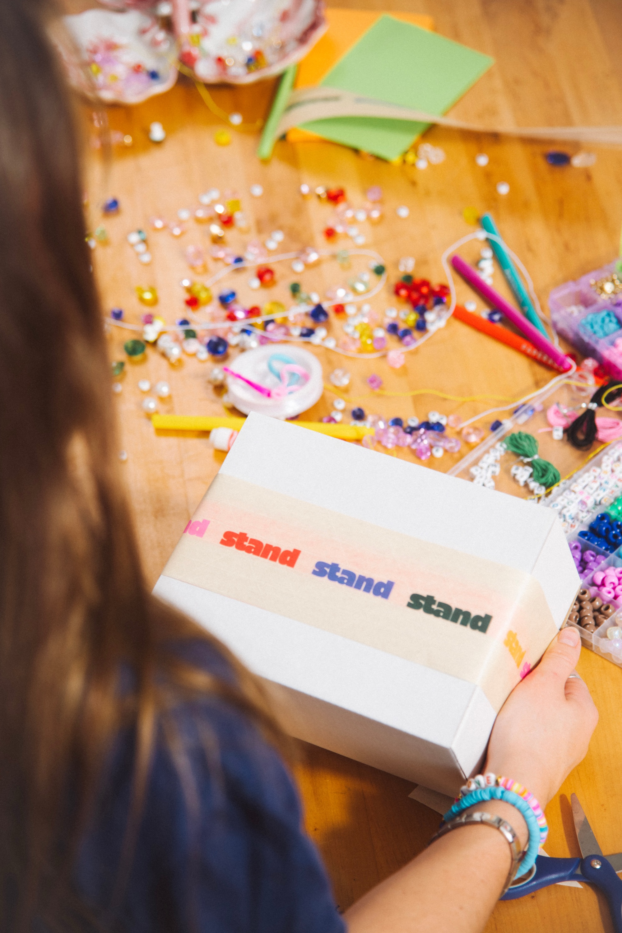 A kid at a craft table covered in beads, holding a white box wrapped in STAND tape